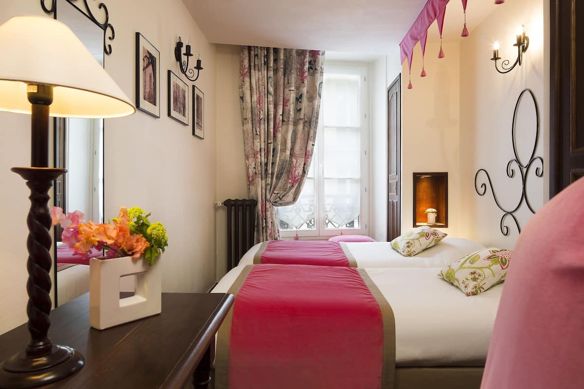 A hotel room with a bed, a bedside table with a lamp, a window with curtains, and wall-mounted lights.