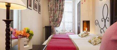 A hotel room with a bed, a bedside table with a lamp, a window with curtains, and wall-mounted lights.