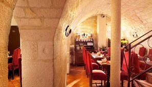 Un comedor con paredes de piedra, sillas tapizadas en rojo y una mesa de madera, con una araña y una escalera.