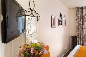 A hotel room with a bed, a television, a mirror, and a floral arrangement.