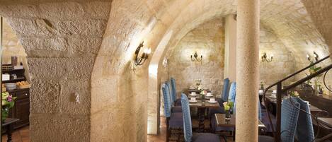 A stone-walled dining area with high tables and chairs, a staircase, and wall-mounted lighting.