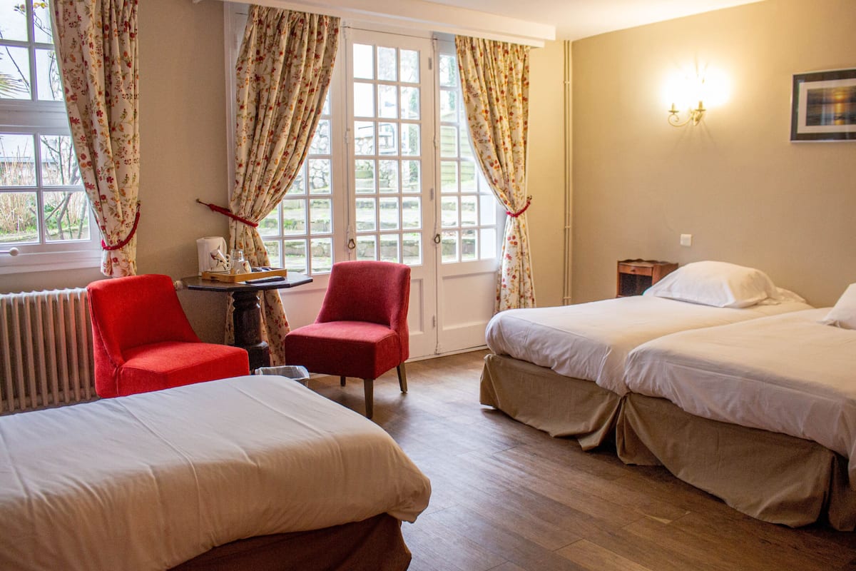 Une chambre d’hôtel avec deux lits, une chaise rouge, une petite table avec une tasse et une fenêtre avec des rideaux.