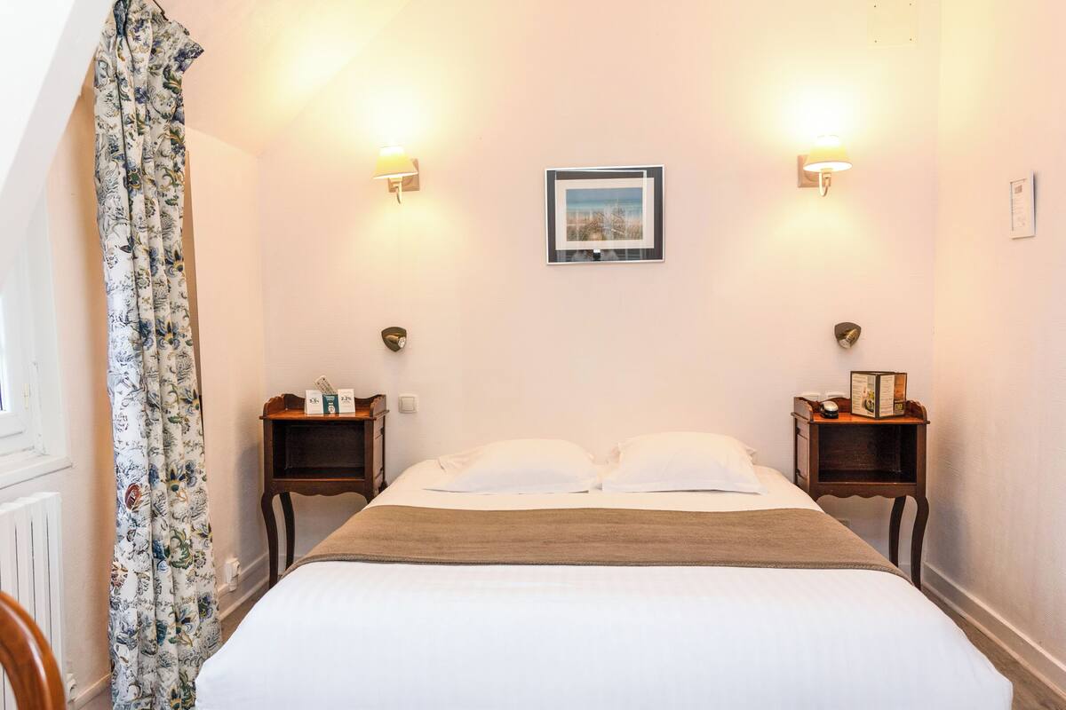 Une chambre à coucher comprenant un lit, deux tables de chevet, une commode, un tableau accroché au mur et un rideau.