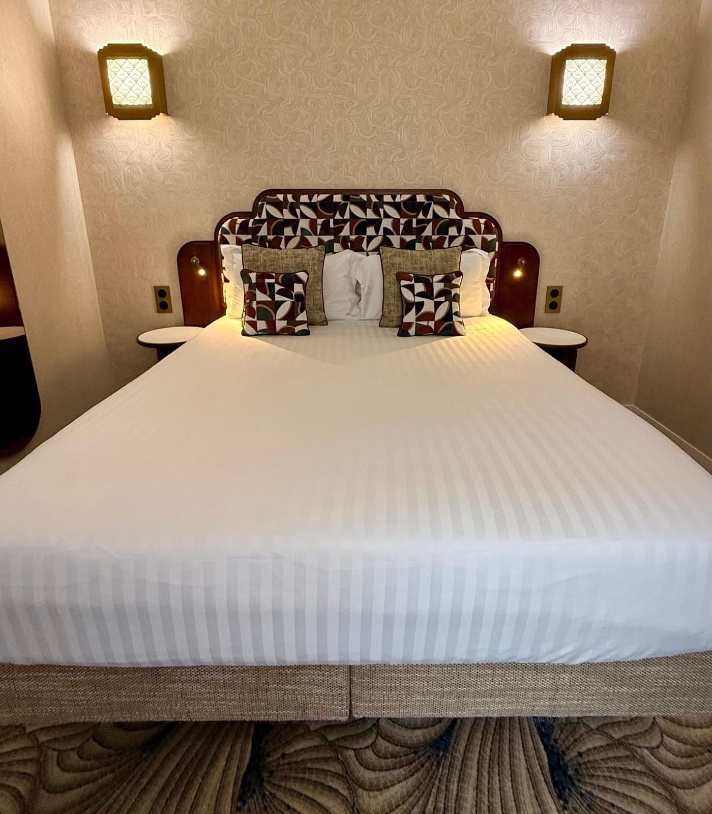 Una habitación de hotel con una cama grande, cabecera estampada, dos mesitas de noche y lámparas de pared.
