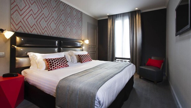Een hotelkamer met een groot bed, een rode bijzettafel, een grijze fauteuil en een behang met een patroon.