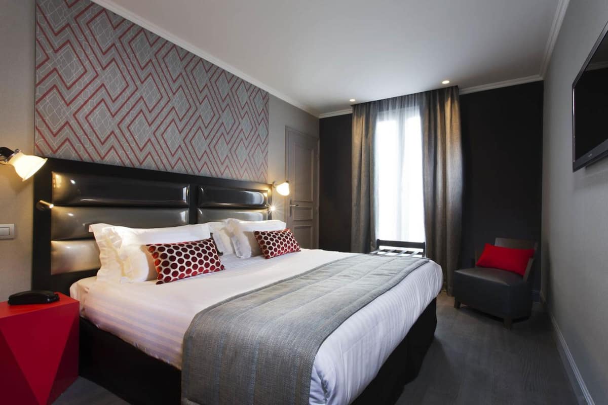 Ein Hotelzimmer mit einem großen Bett, einem roten Beistelltisch, einem grauen Sessel und einer Wand mit Muster.