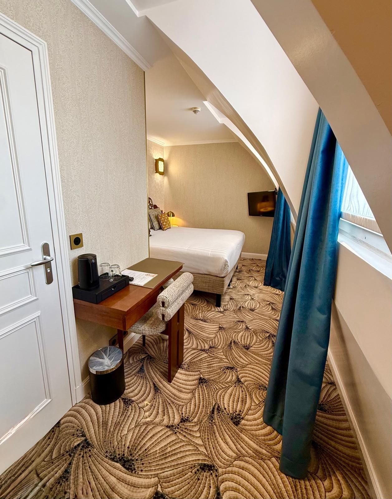 A hotel room with a bed, a desk, a chair, a trash bin, a wall-mounted TV, and a window with curtains.