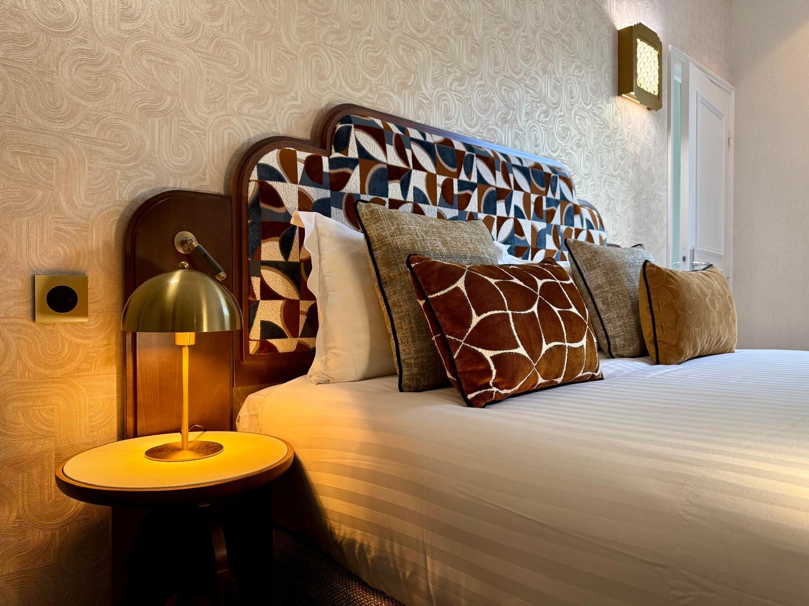 Una habitación de hotel con un cabecero estampado, una lámpara de noche y varias almohadas.