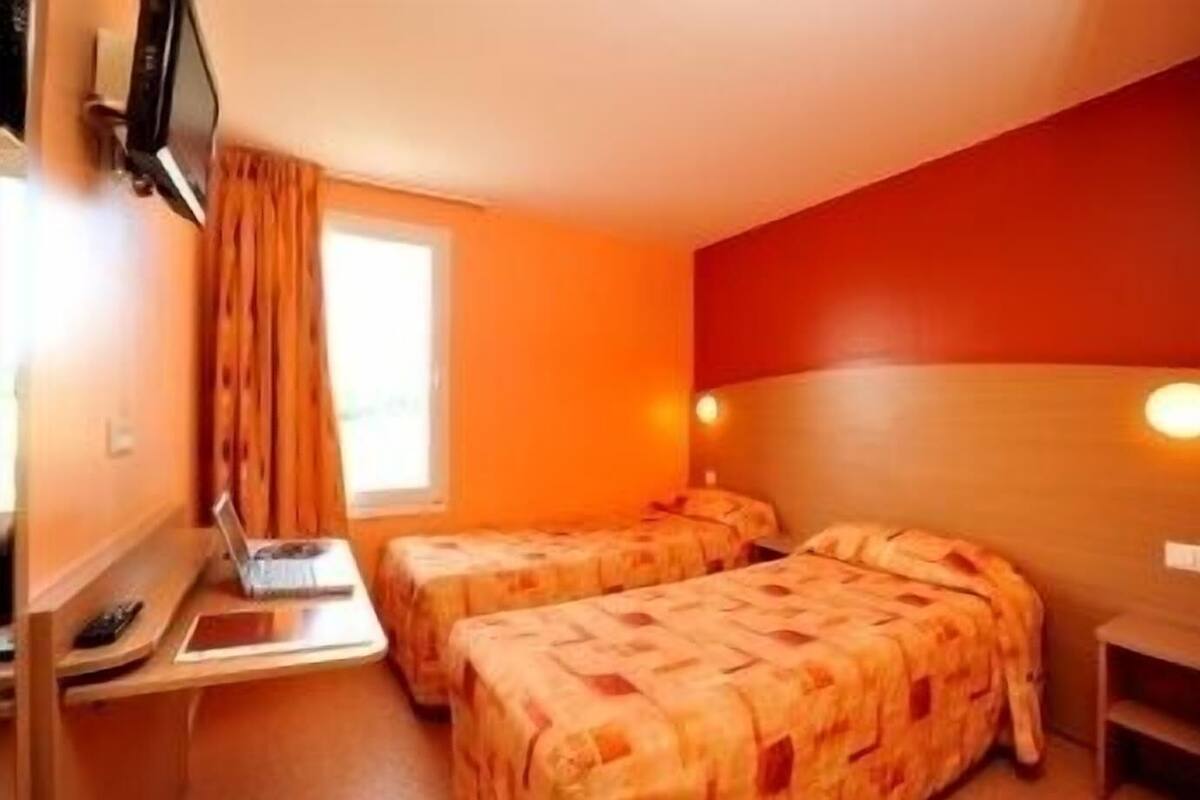 Een hotelkamer met twee bedden, een bureau met een laptop, een televisie en een raam met gordijnen.