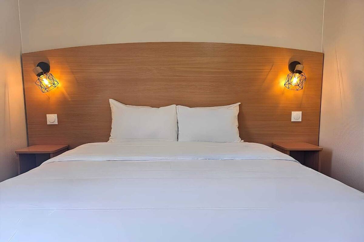Een net opgemaakt bed met witte lakens, een houten hoofdbord en twee bedlampjes.
