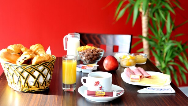Een ontbijttafel met croissants, sinaasappelsap, een koffiekop, een kom cereal, een boterham met ham en een schaal fruit.