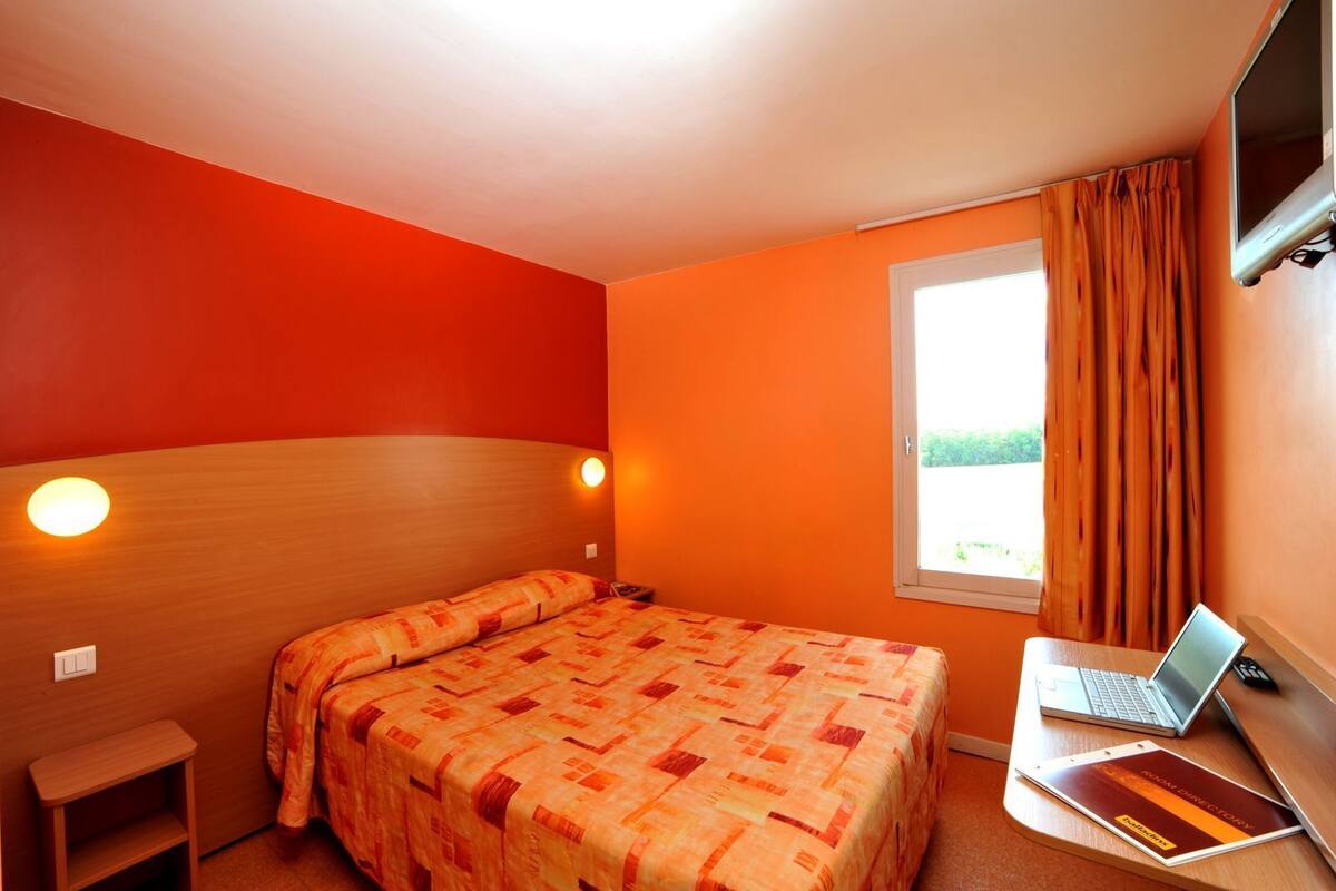 Een hotelkamer met een bed, een bureau met een laptop, een televisie en een raam met gordijnen.