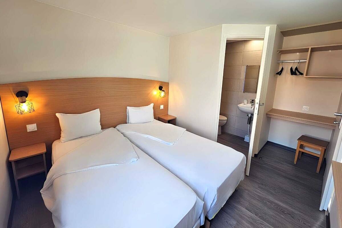 Hotelkamer met twee bedden, een houten hoofdbord, bedlampen, een klein tafeltje, een badkamer met een wastafel en spiegel, en een houten plank met hangende items.