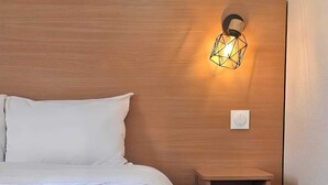 Een net opgemaakt bed met witte lakens, een houten hoofdbord, een klein houten nachtkastje en een wandlamp.