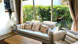 Un salon accueillant avec un canapé, une table basse en bois et une table d’appoint avec une lampe. Une fenêtre offre une vue sur un paysage verdoyant.
