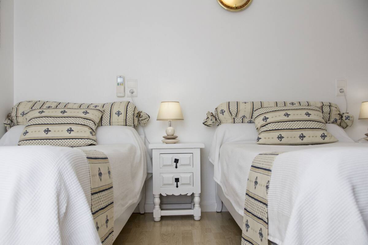 Habitación doble con camas con cabeceros a juego, una mesita de noche con lámpara y un pequeño aparador blanco con cajones.