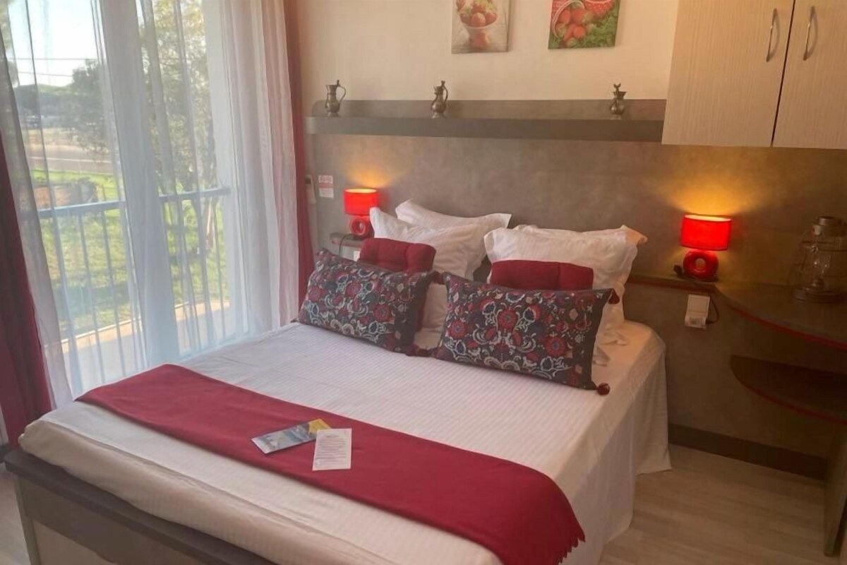 A neatly made bed with a red blanket, two bedside lamps, and a view of the outdoors through a window.