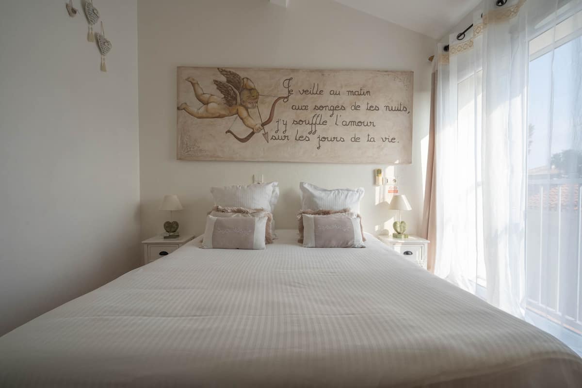 Una cama bien hecha con un adorno grande en la pared, dos mesitas de noche con lámparas y una ventana con cortinas traslúcidas.