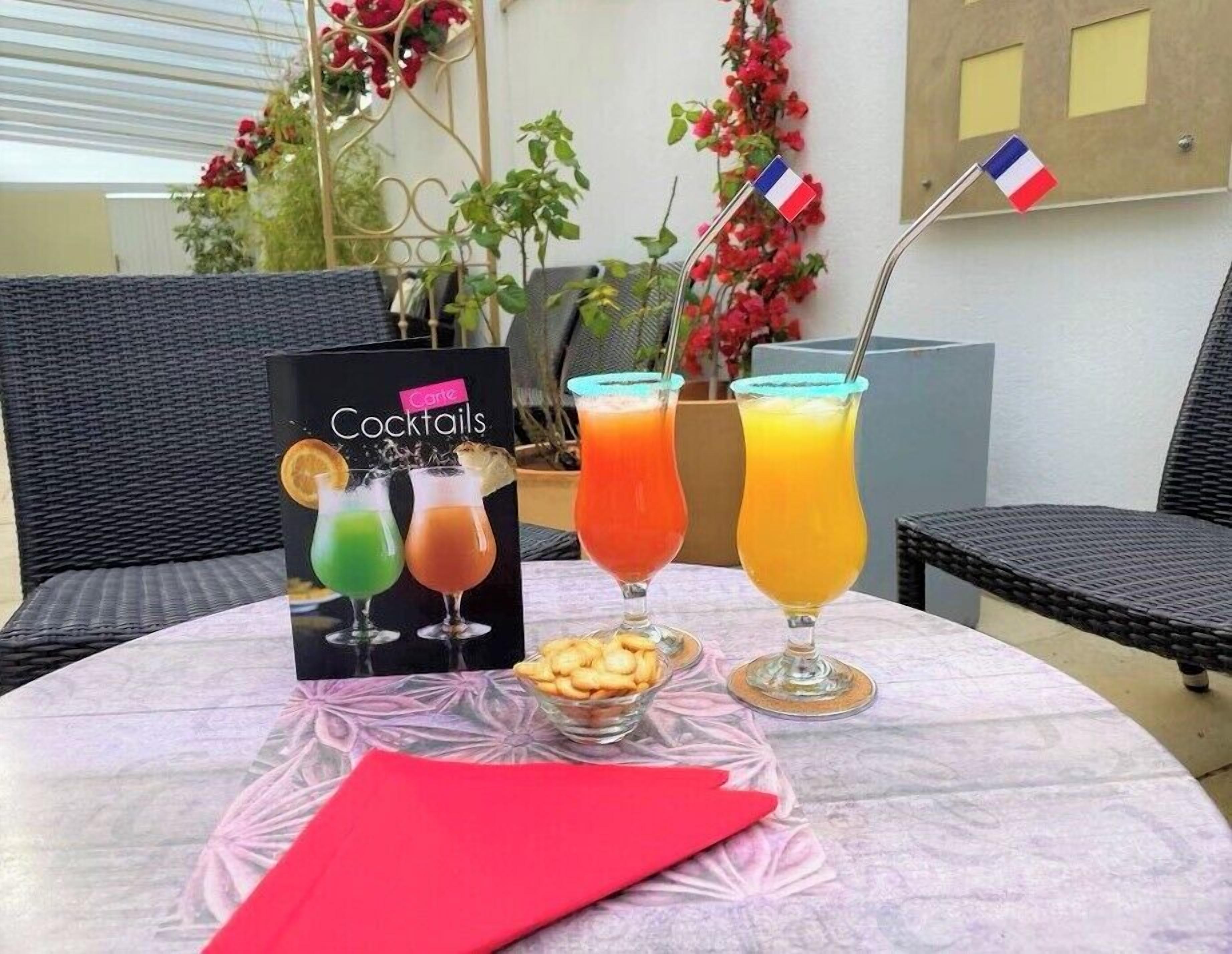 A table with a cocktail menu, three drinks, a bowl of snacks, and a French flag.