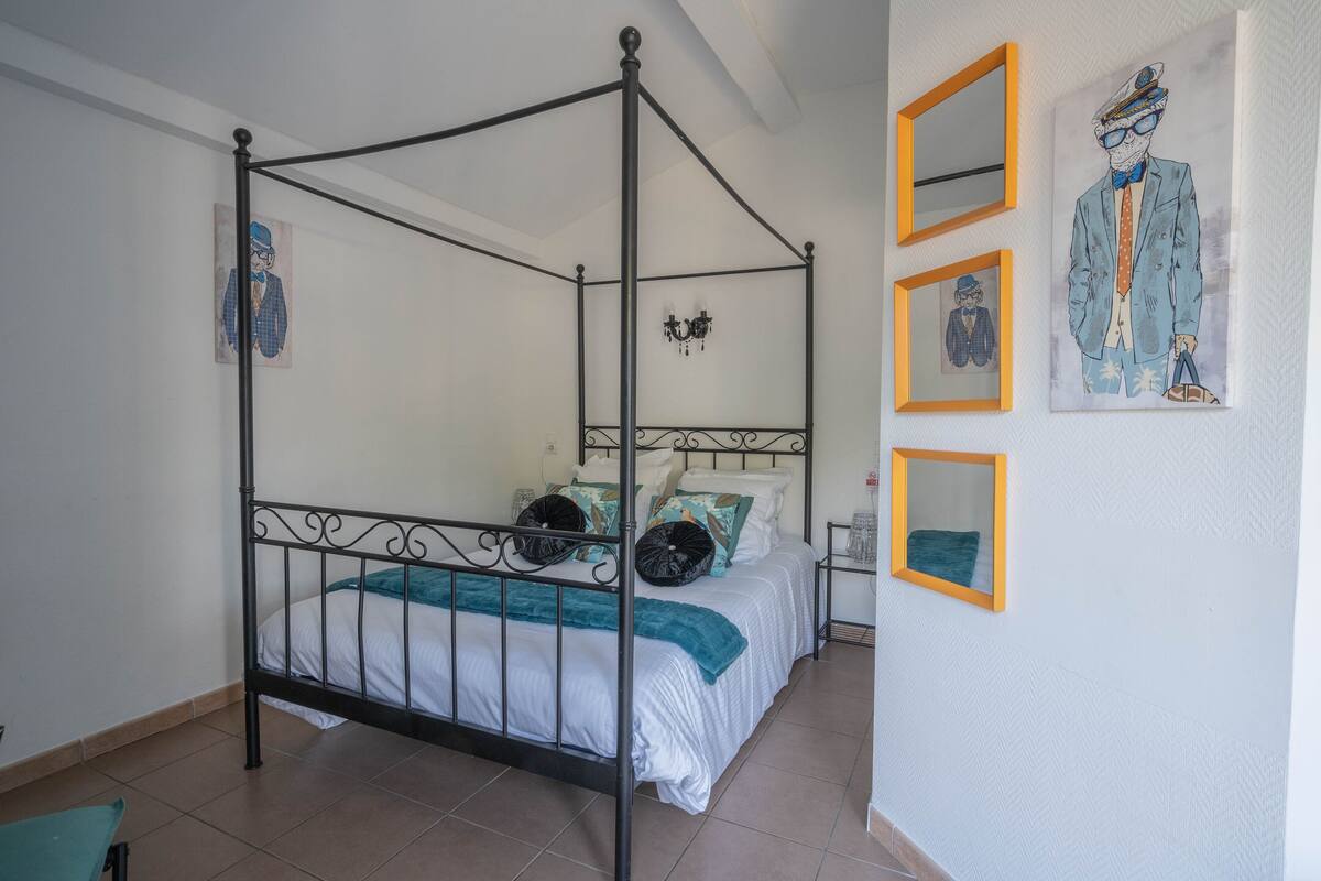Una cama con dosel con ropa de cama blanca y detalles color esmeralda, una mesita de noche pequeña con una lámpara, y obras de arte en las paredes.