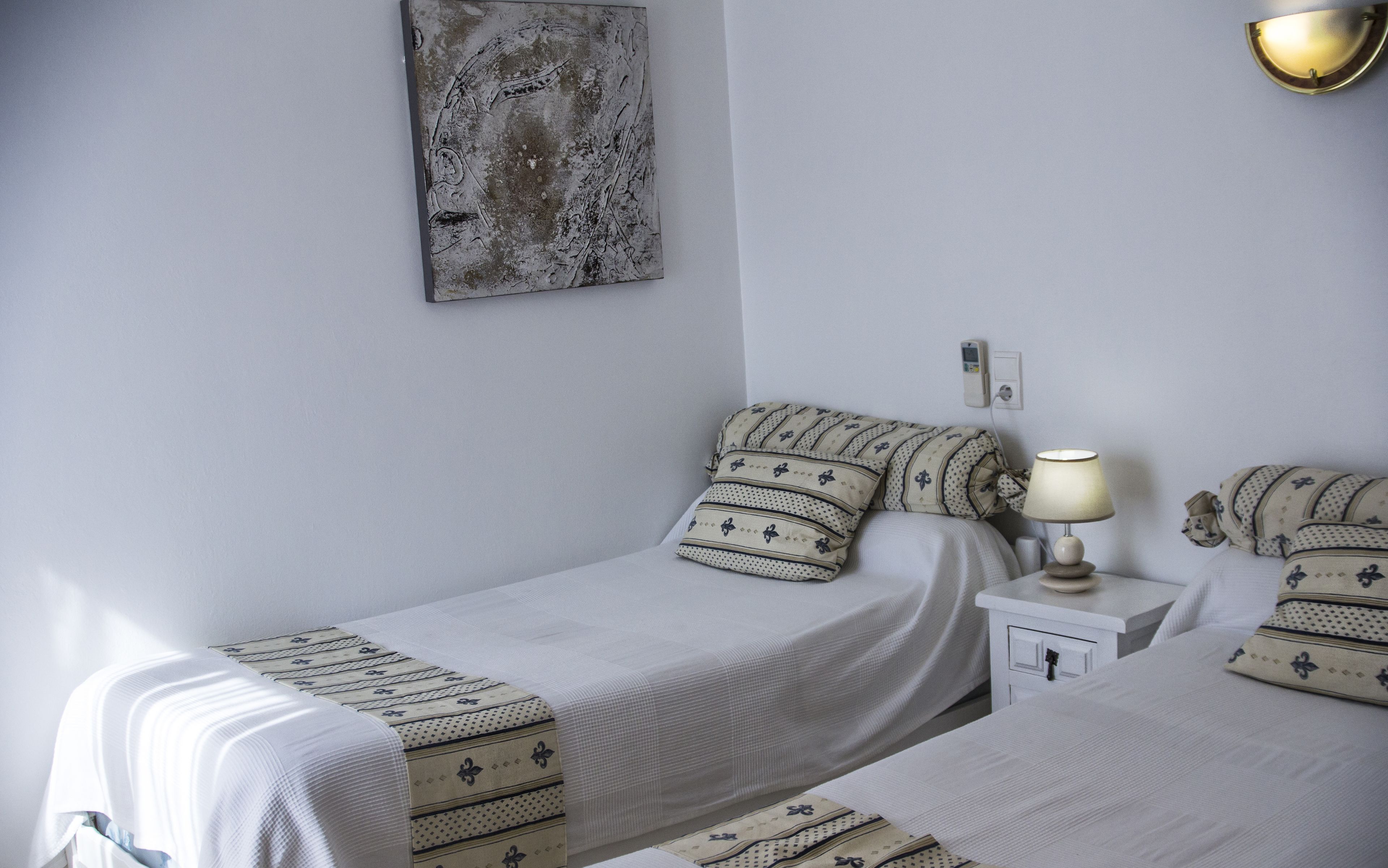 Un dormitorio con dos camas, una mesita de noche con lámpara, una luz empotrada en la pared y un cuadro.