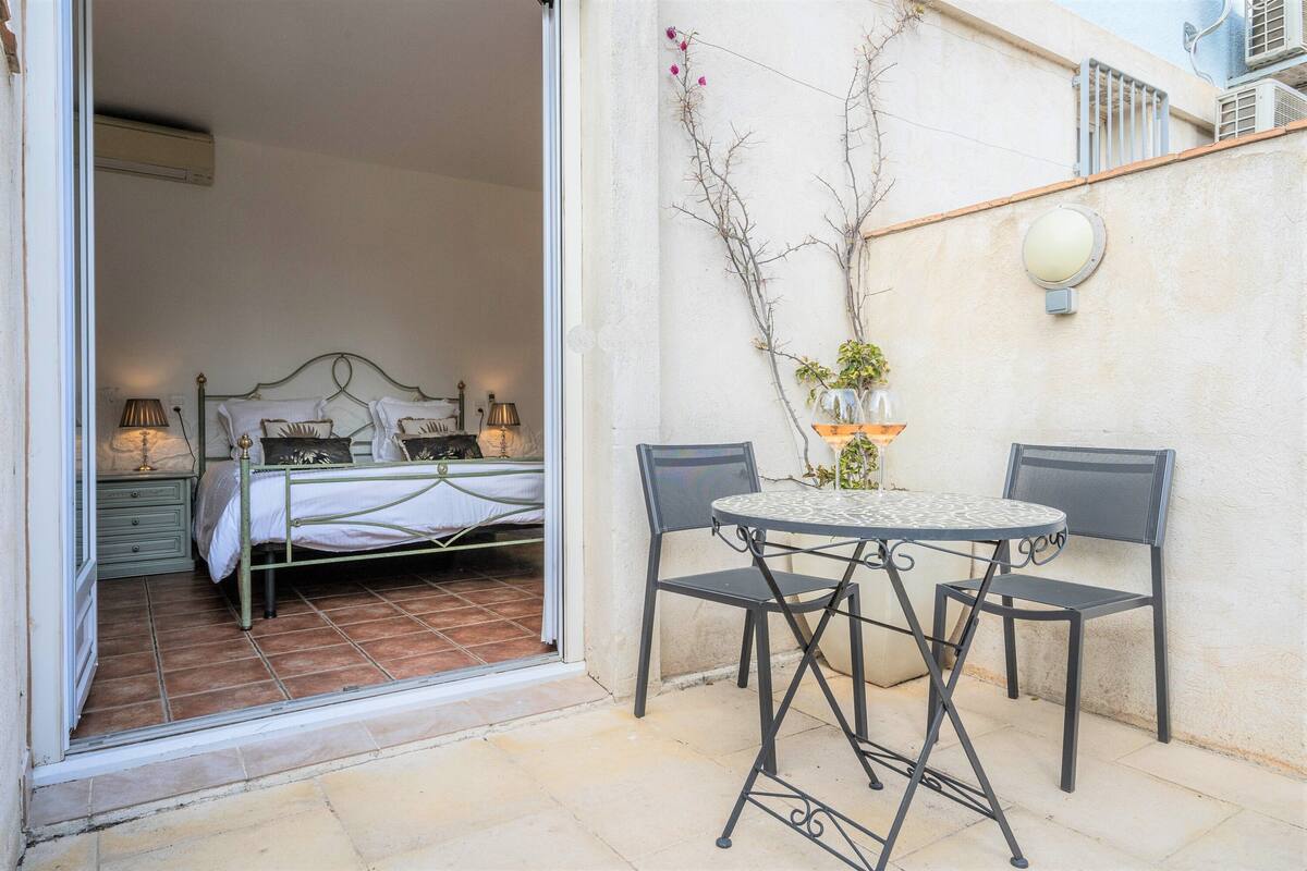 Un dormitorio con cama, mesitas de noche y ventana. Cuenta con un pequeño patio con mesa y sillas, una planta en maceta y un aplique de pared.