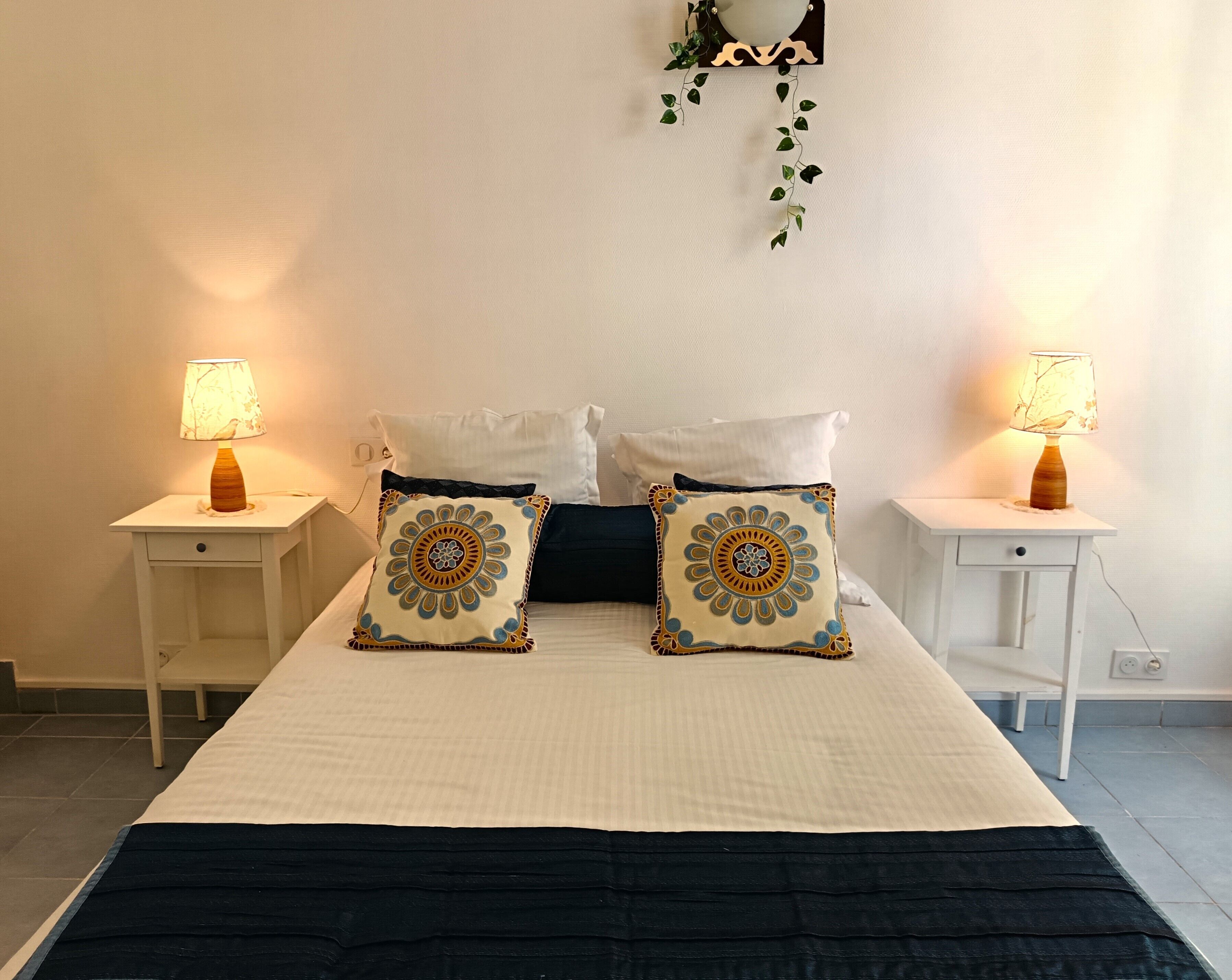 A neatly made bed with two decorative pillows, two bedside tables with lamps, and a wall-mounted plant.