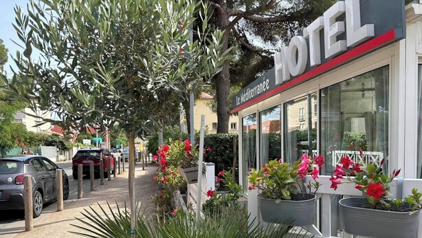 Un hôtel à l’ambiance méditerranéenne, avec une terrasse agrémentée de plantes en pot et donnant sur la rue, où l’on peut voir des voitures garées.