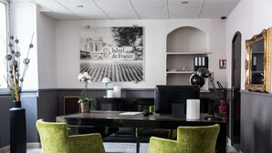 Een hotel receptie met een balie, twee groene stoelen, een computer, een vaas met bloemen, een groot ingelijst schilderij en een spiegel.