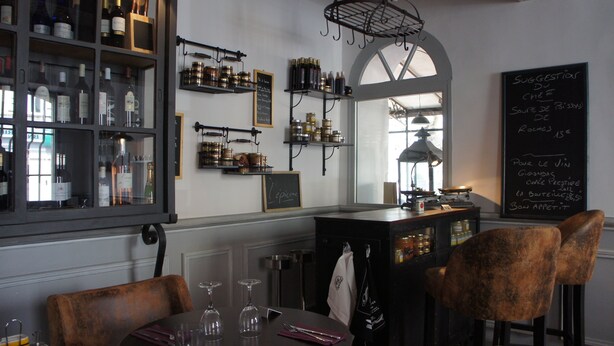 Een restaurantinterieur met een bar, planken met flessen en potten, een krijtbordmenu en een raam met uitzicht.