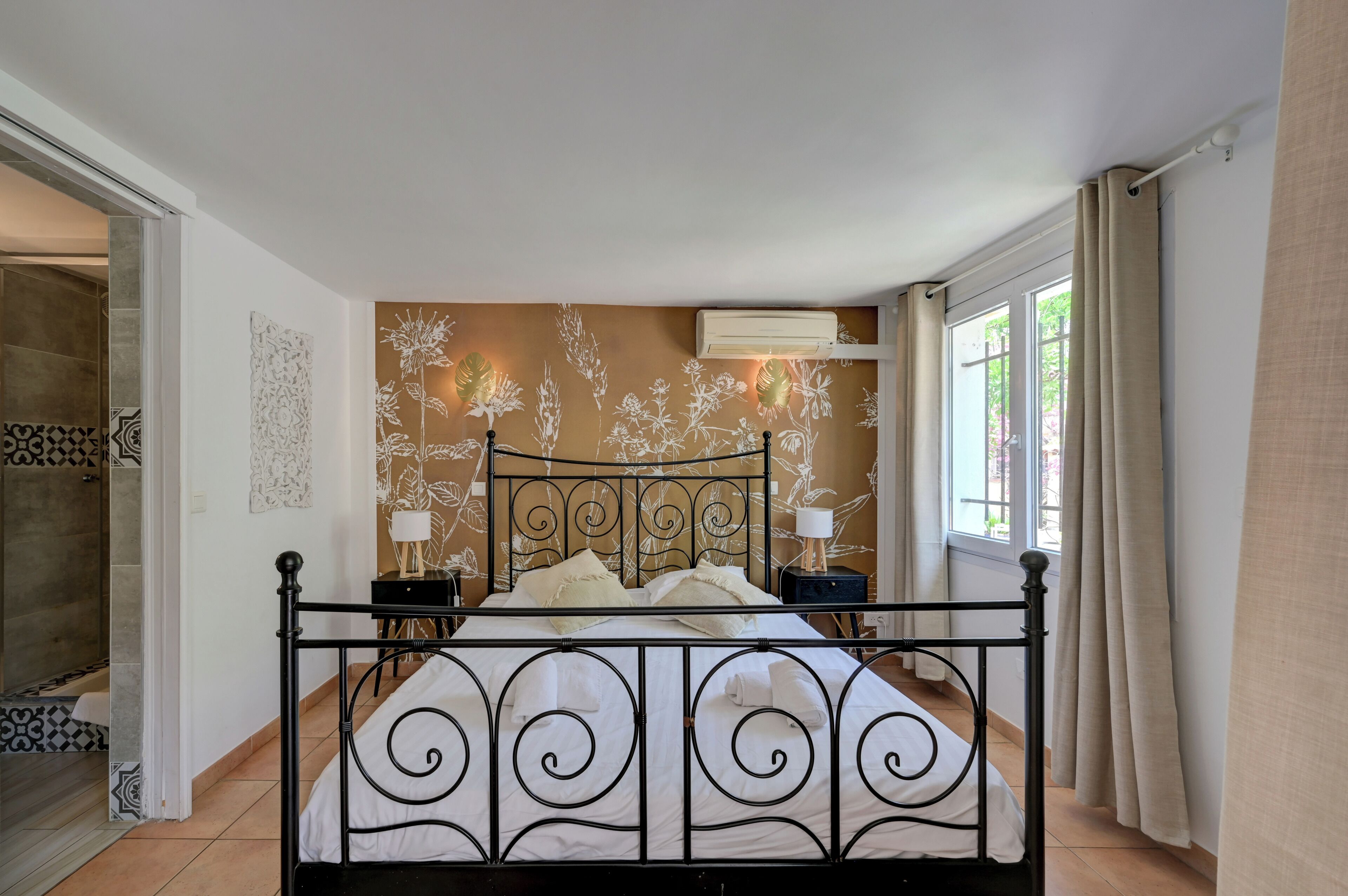 Une chambre à coucher avec un lit en métal, une table de chevet avec une lampe, un climatiseur fixé au mur et une fenêtre avec des rideaux.