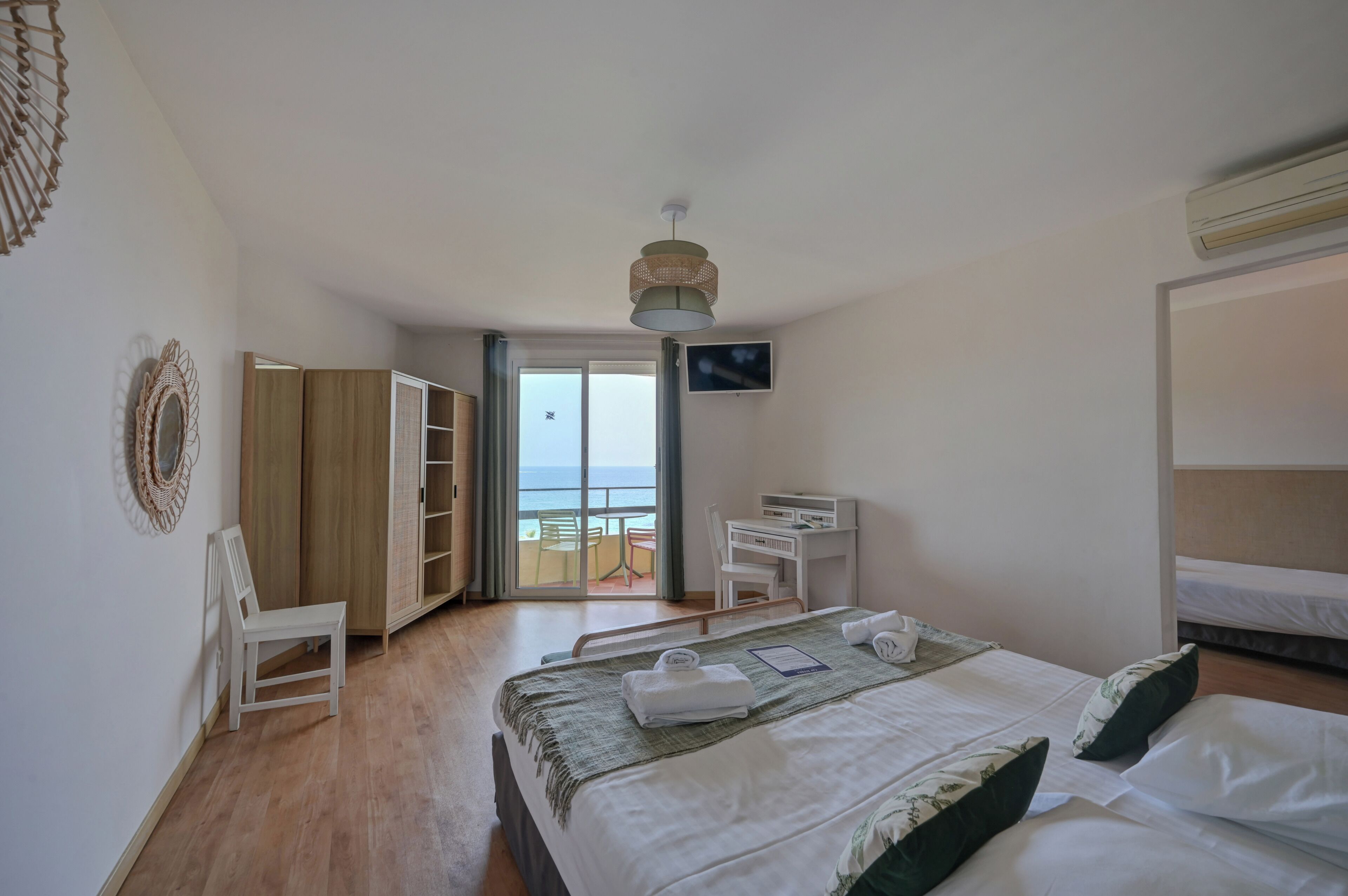A hotel room with a bed, a desk, a chair, a TV, and a balcony with a view of the sea.