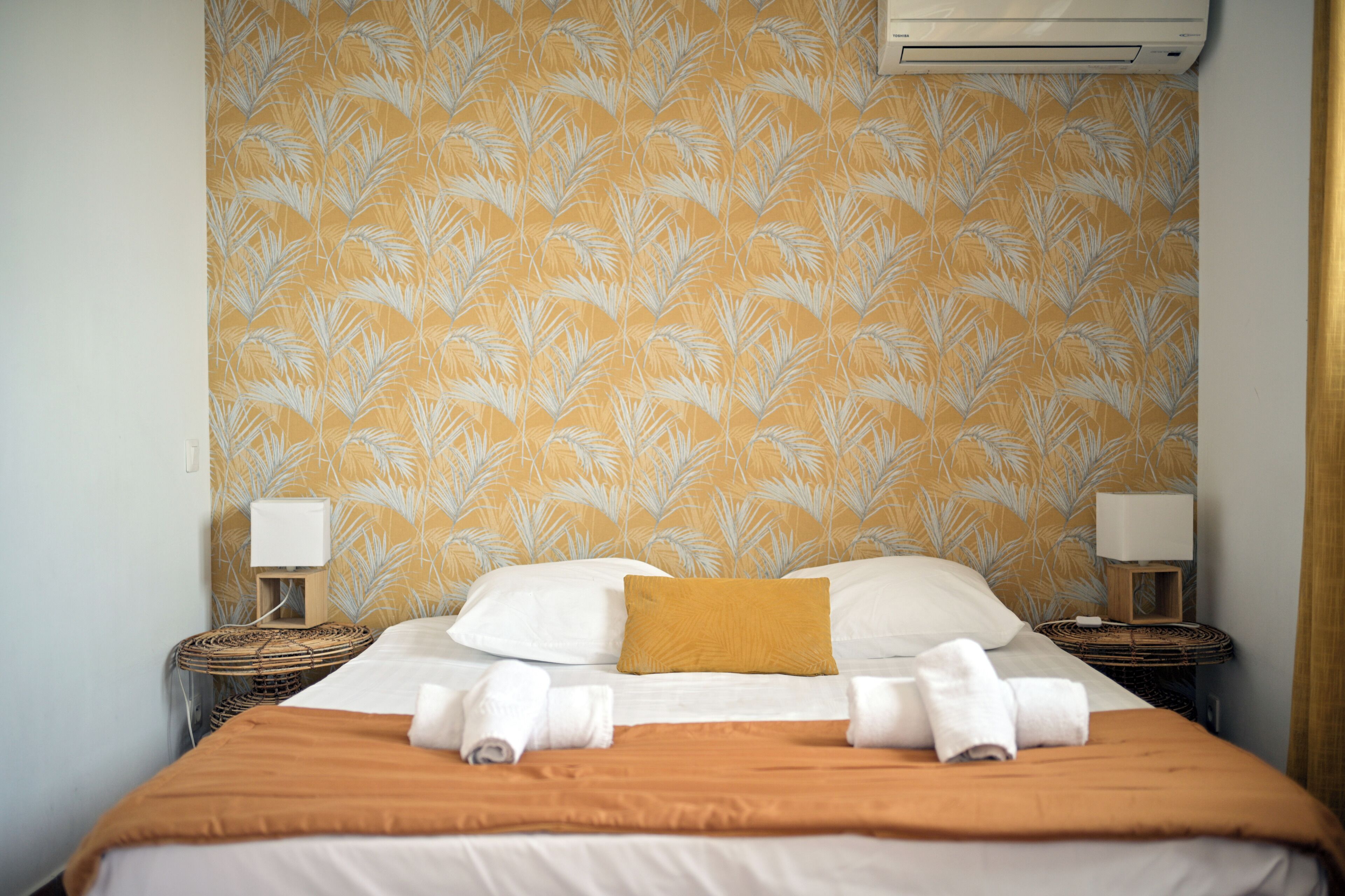 Habitación de hotel con papel tapiz estampado, una cama con ropa de cama blanca y marrón, dos mesitas de noche con lámparas y un aire acondicionado de pared.