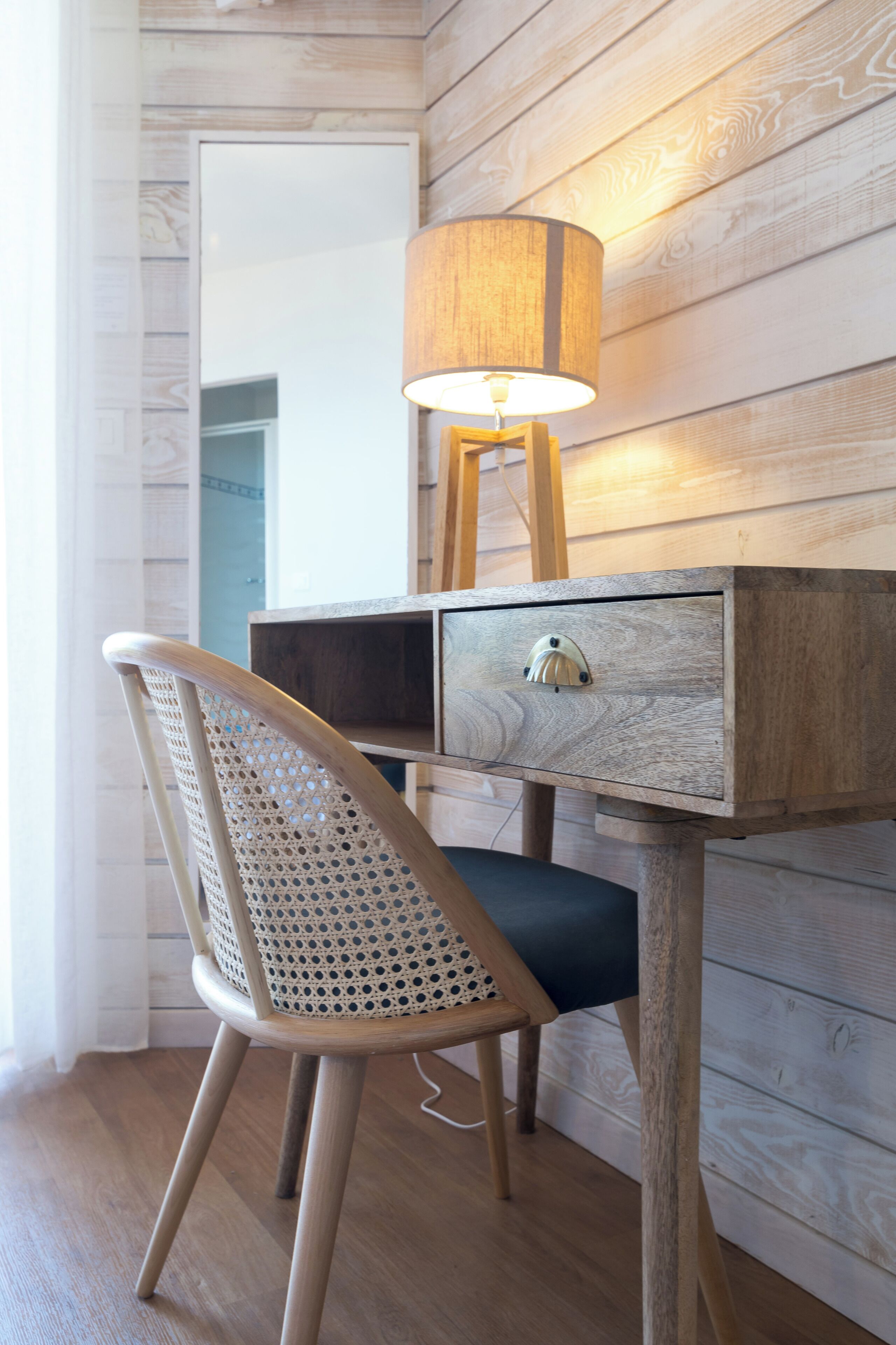 A wooden desk with a lamp, a chair with a woven backrest, and a drawer.
