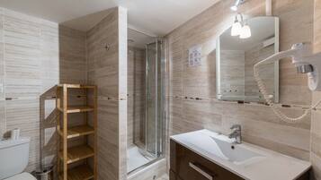 Un baño moderno con ducha de vidrio, estante de madera, lavamanos blanco y espejo con secador de pelo.