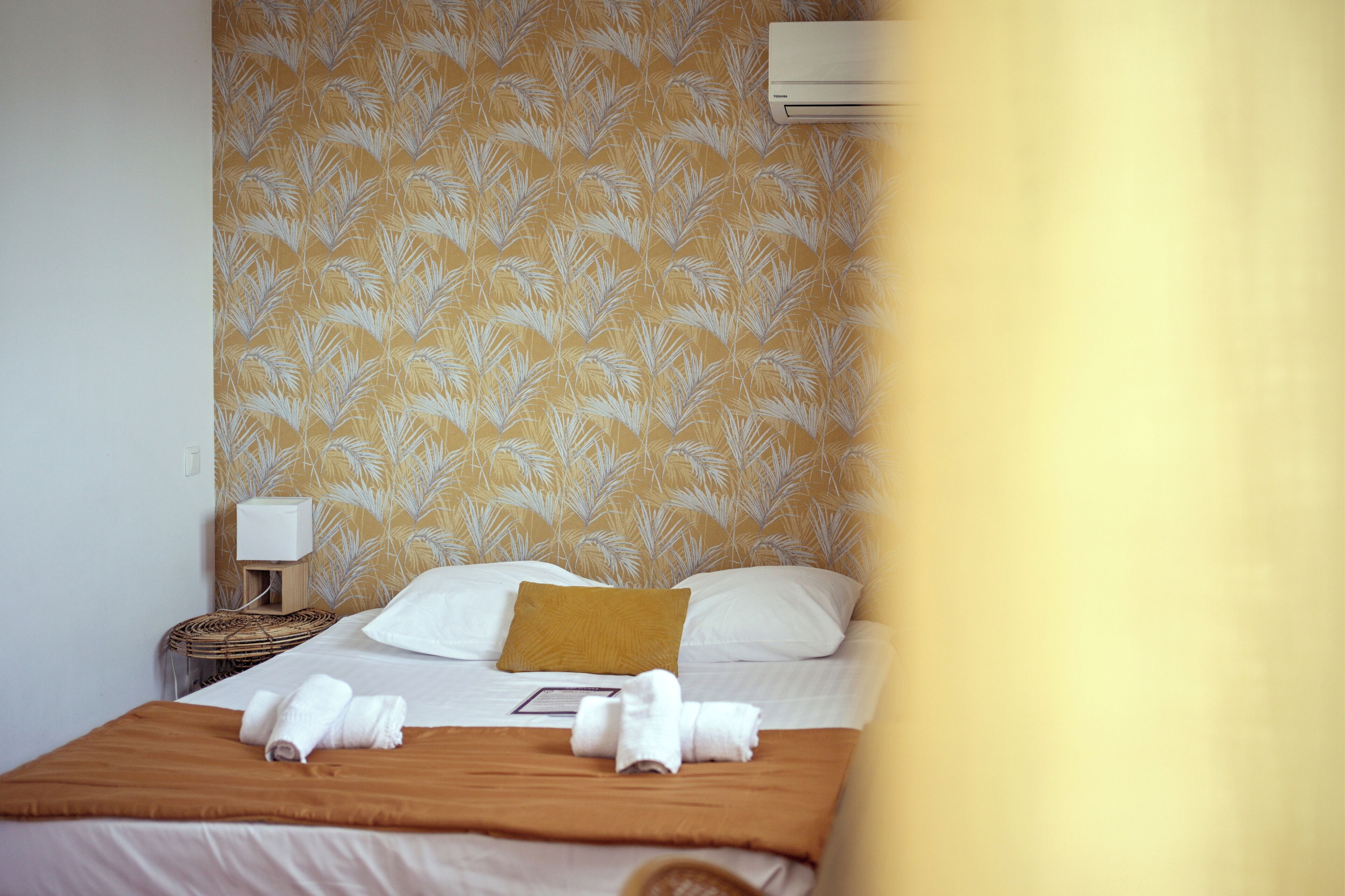 Een keurig opgemaakt bed met witte lakens, een bruine plaid en twee opgerolde handdoeken op het bed.