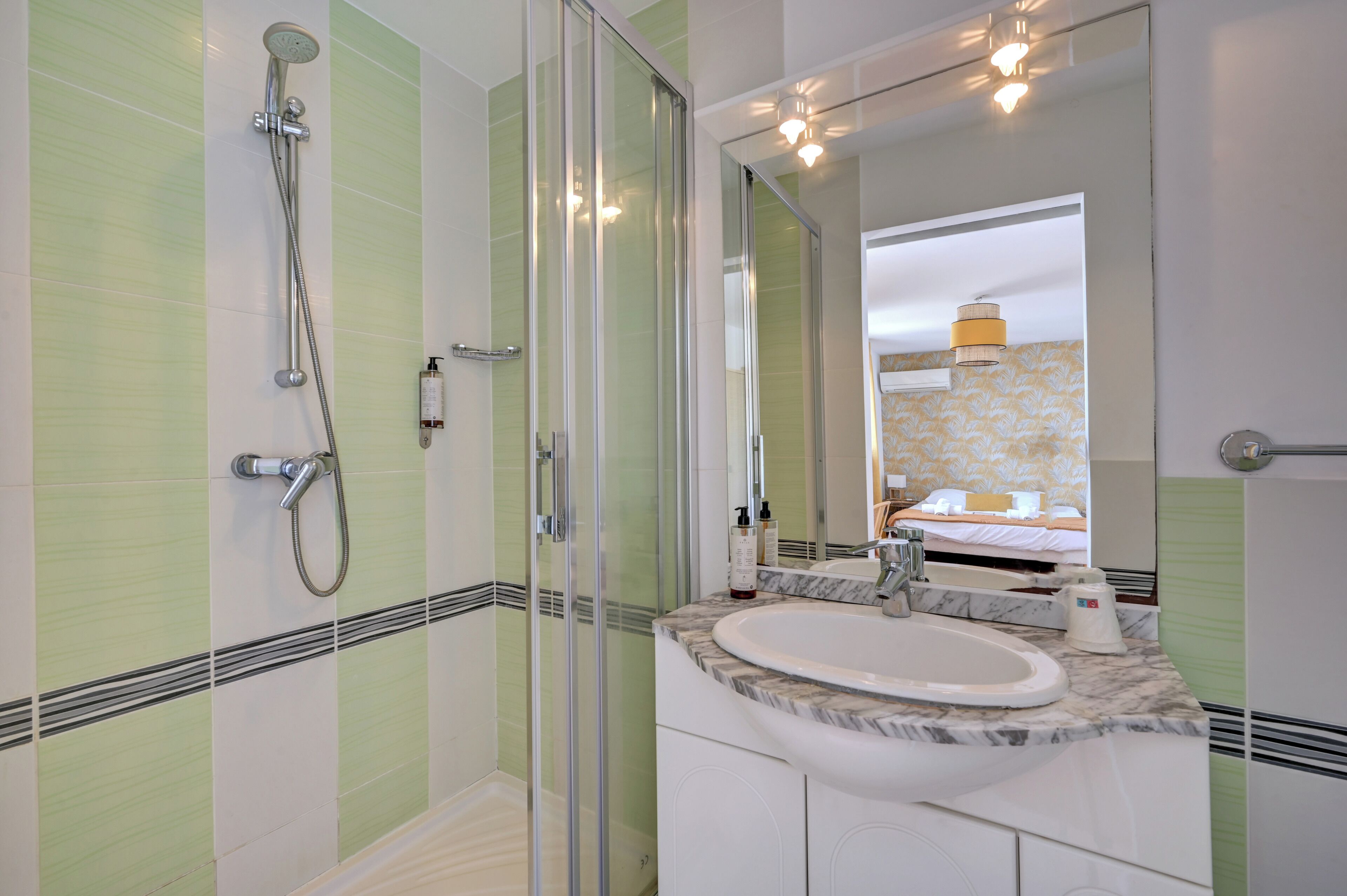 Un baño con cabina de ducha de vidrio, un lavamanos con encimera de mármol y un espejo que refleja una cama con una lámpara amarilla.