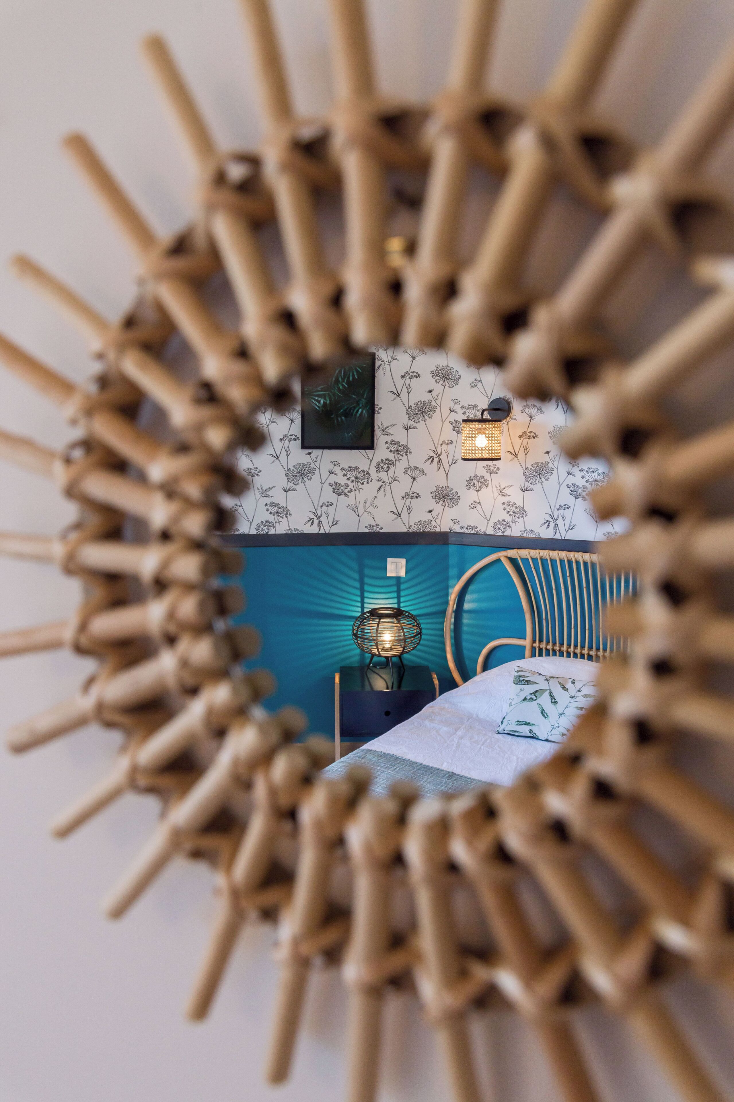 A circular mirror with a bamboo frame, a wall with floral wallpaper, a blue accent wall, and a bed with a white bedspread.