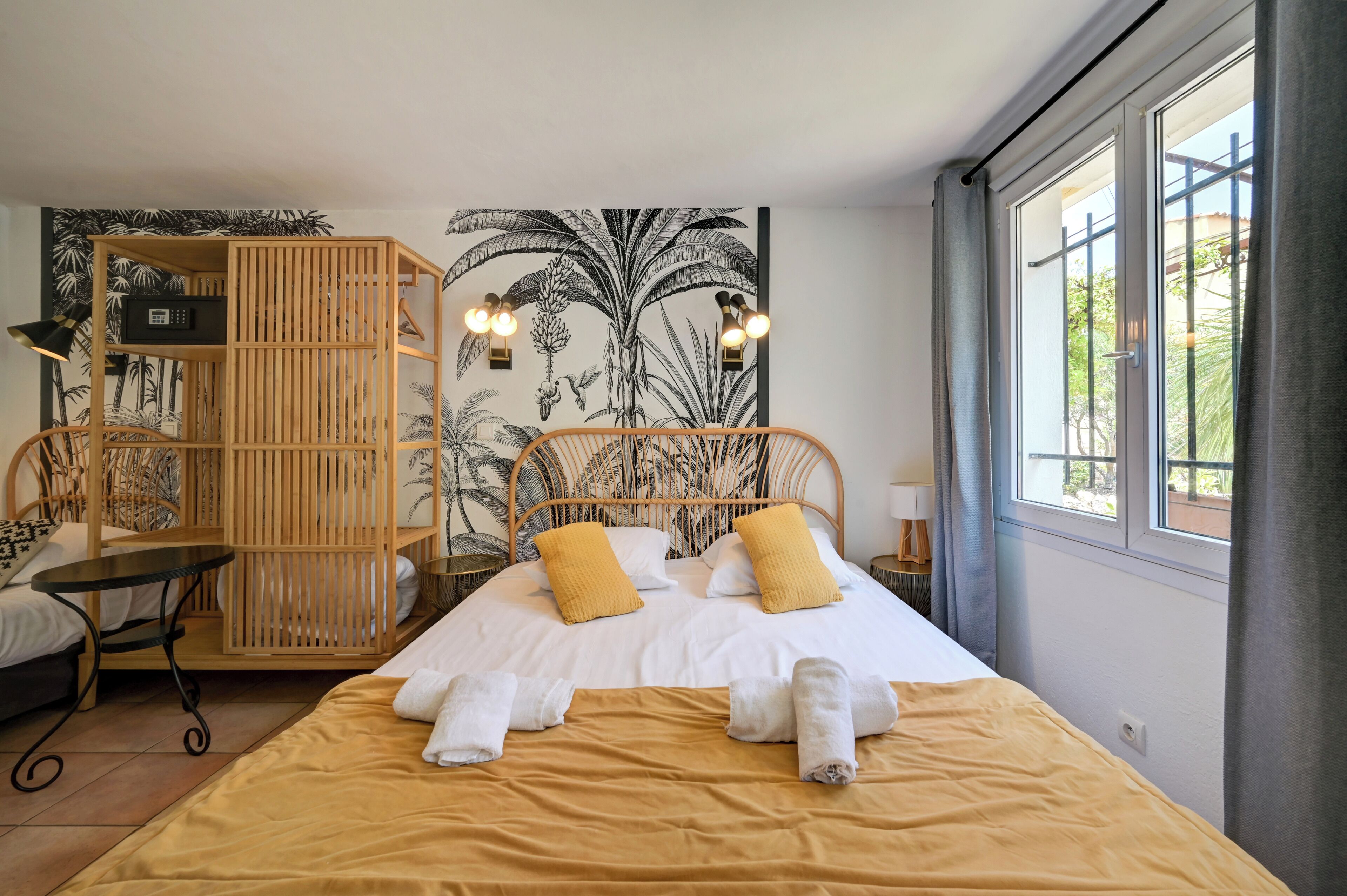 A bedroom with a bed, a wooden wardrobe, a small table, and a wall with a tropical mural.