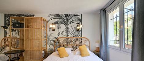 A bedroom with a bed, a wooden wardrobe, a small table, and a wall with a tropical mural.