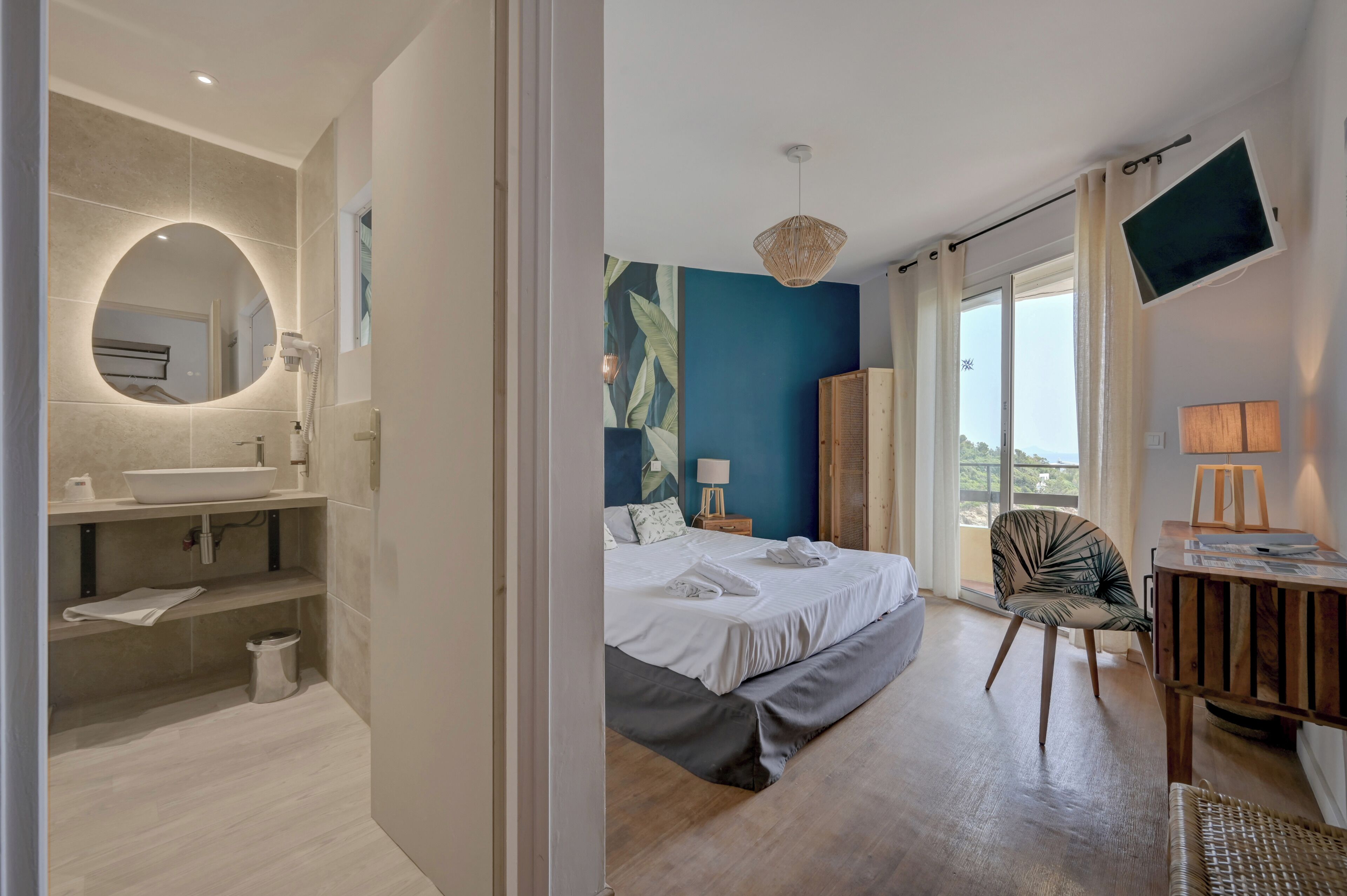 A modern hotel room with a large bed, a bathroom with a round mirror, a TV, and a view of the outdoors.