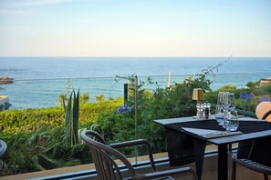 A dining table with chairs, a view of the ocean, and some greenery.