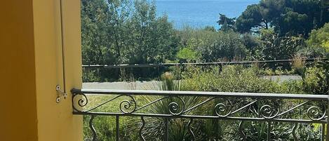 Varanda com mobiliário de vime, guarda-corpo de metal e vista para o mar e vegetação.