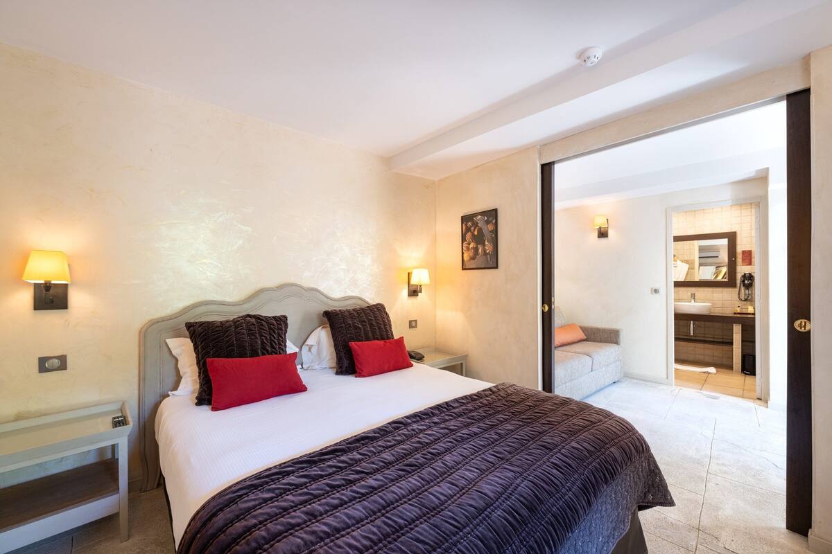 Une chambre avec un grand lit, deux tables de chevet, une commode, une porte donnant sur une salle de bain et un luminaire fixé au mur.