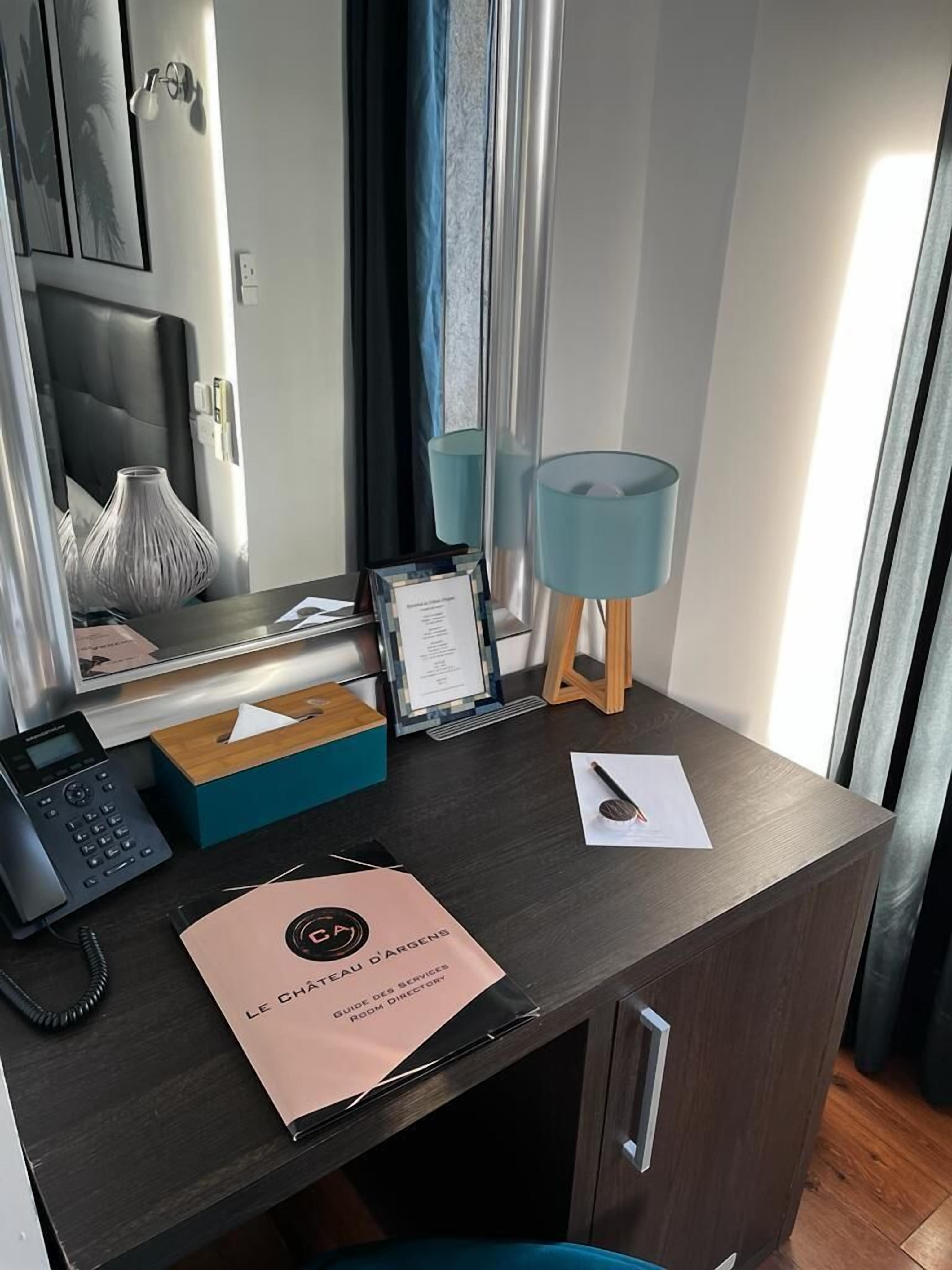 Escritorio de habitación de hotel con un teléfono, una caja de pañuelos, un cartel y una lámpara.