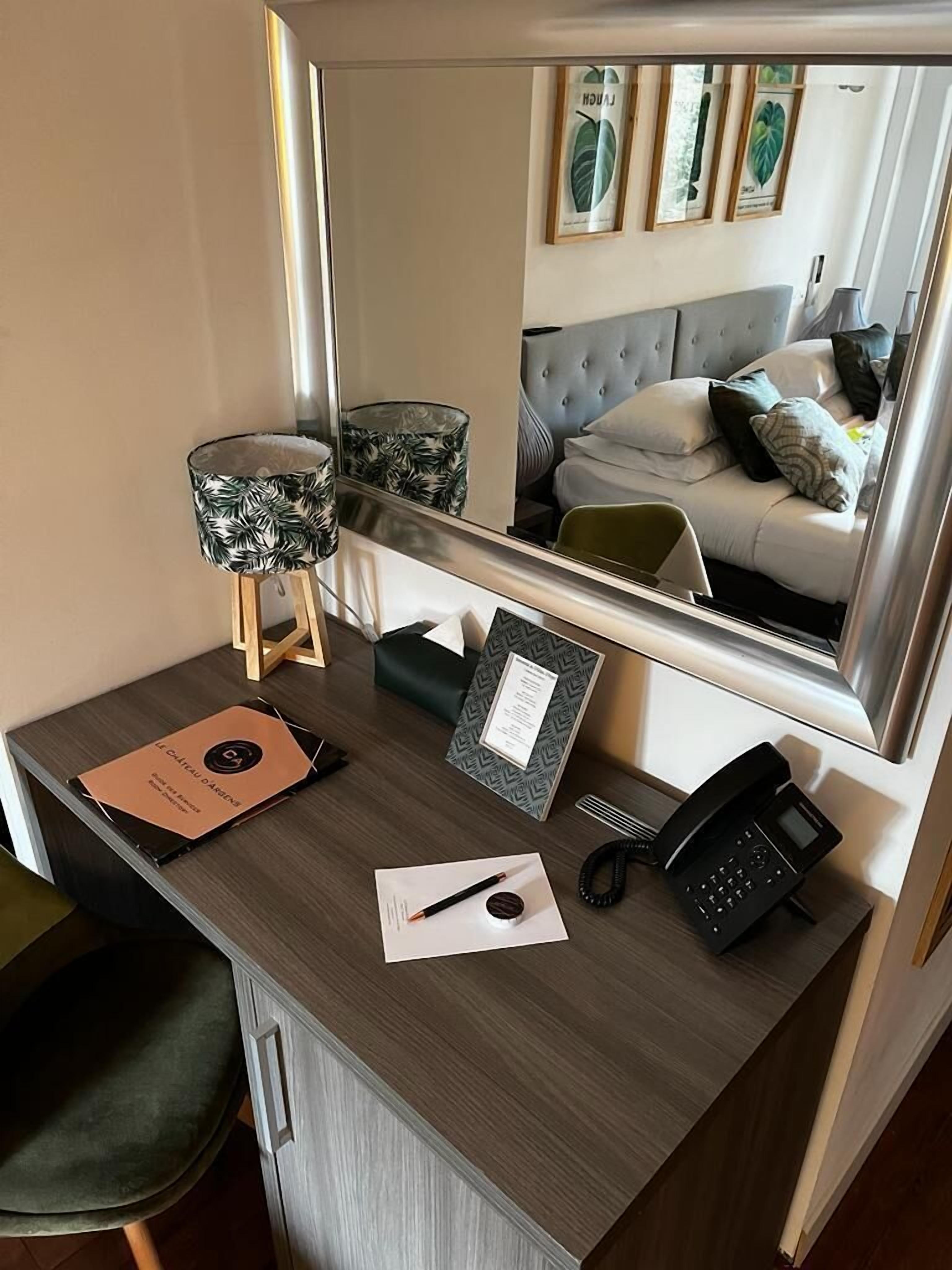 Un escritorio de hotel con un espejo que refleja un dormitorio. El escritorio tiene una lámpara, un teléfono, un cuaderno y una carpeta.