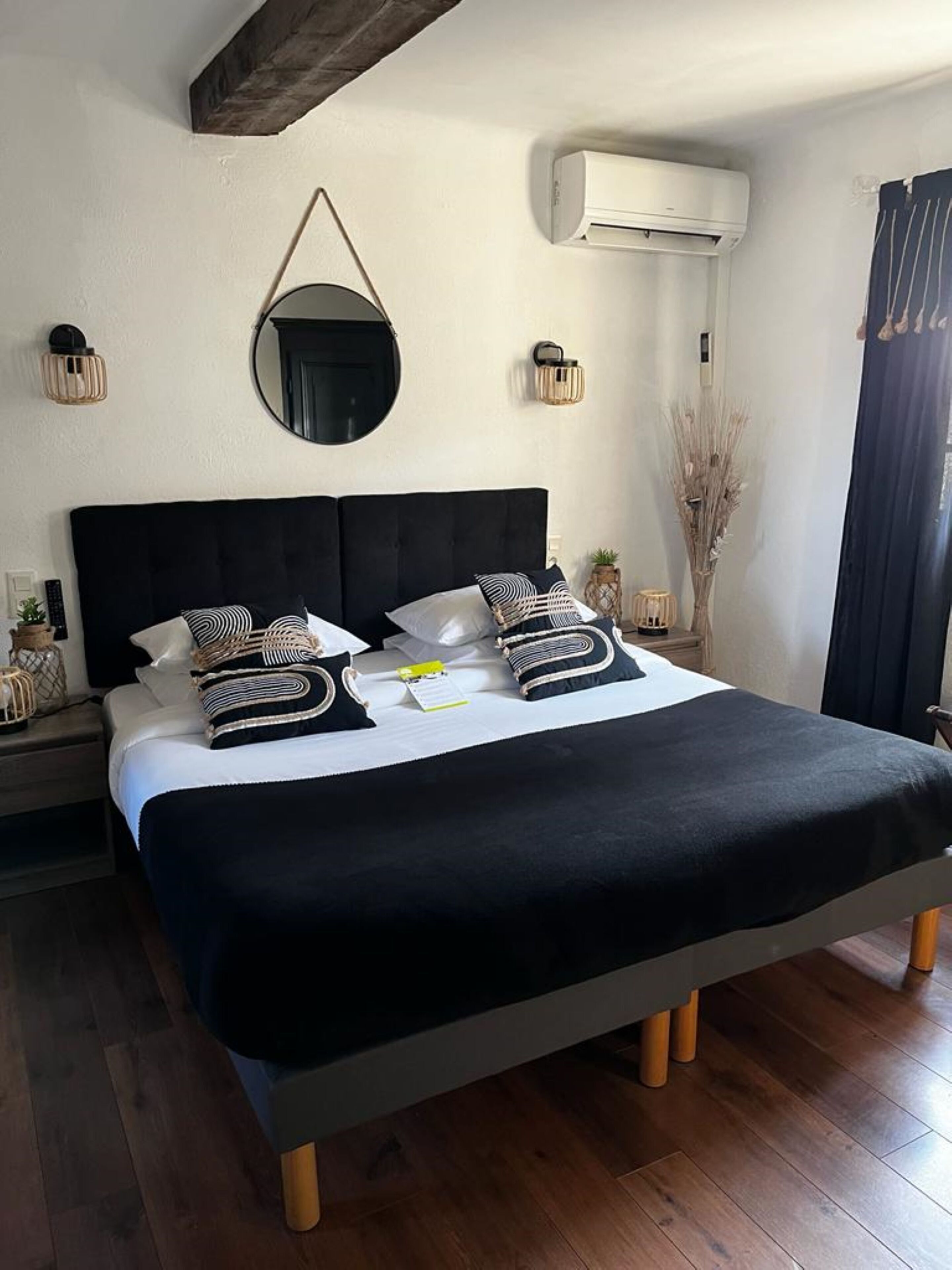 Un letto rifatto con cura, con testiera nera, biancheria bianca e cuscini decorativi. Si nota un condizionatore d'aria a parete e uno specchio rotondo sopra il letto.