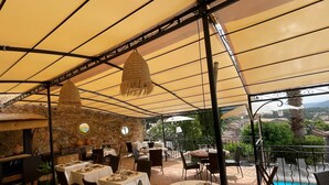 Un espace extérieur aménagé pour les repas, avec des tables et des chaises, un mur en pierre et une vue sur une piscine et un jardin.