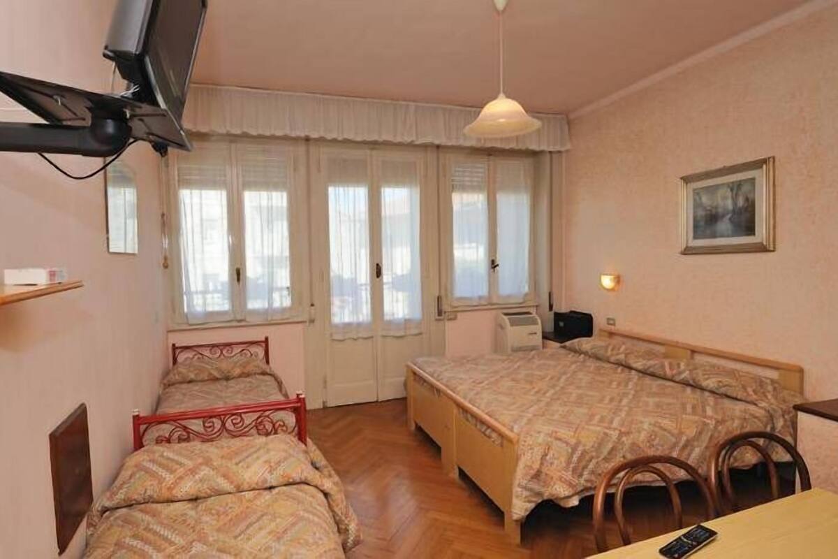 A bedroom with two beds, a TV mounted on the wall, a desk with a chair, and a window with curtains.