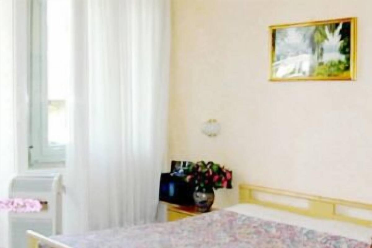 A bedroom with a bed, a window with curtains, a bedside table with a vase of flowers, and a painting on the wall.